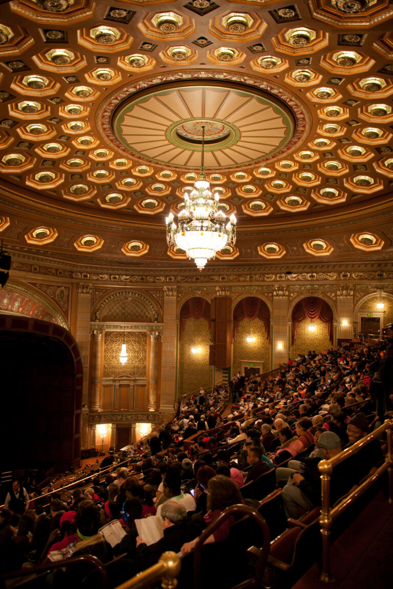 an audience sits in a dimly lit theater under an ornate ceiling with a glowing chandelier